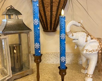 May include: Two blue LED candles with white snowflake designs are displayed in bronze-colored candle holders. A remote control sits in front of one candle. A decorative lantern and a white elephant statue are in the background.
