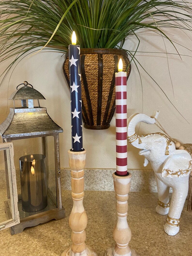 May include: Two decorative electric candles with wooden bases. One candle is navy blue with white stars, the other is red and white striped. The candles are lit and placed on a countertop with other decorative items.
