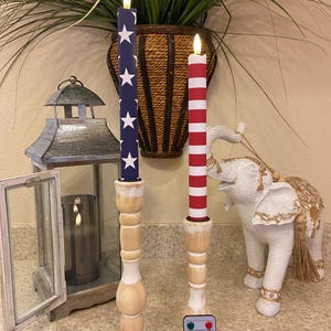 May include: Two battery-operated patriotic candles with a remote. One candle has a blue background with white stars, the other has red and white stripes. Both candles sit on wooden candle holders.
