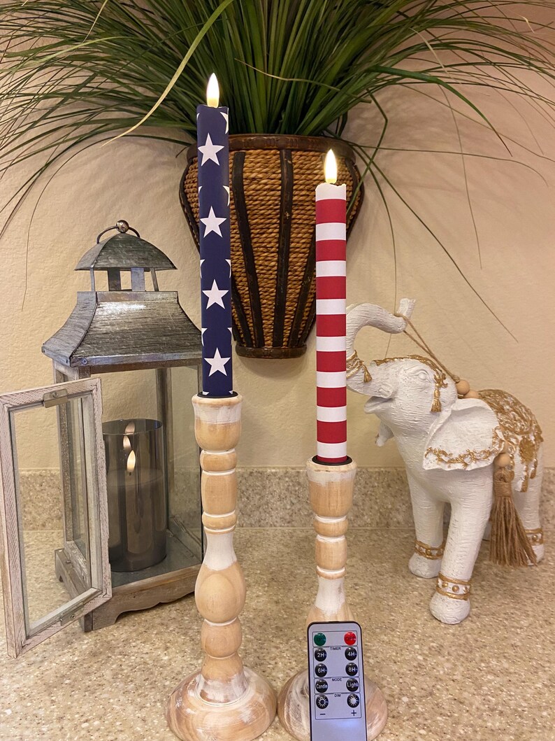 May include: Two battery-operated patriotic candles with a remote. One candle has a blue background with white stars, the other has red and white stripes. Both candles sit on wooden candle holders.