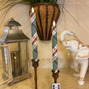 May include: Two battery-operated taper candles with a green, white, and red plaid pattern, set in bronze candle holders. A remote control sits in front of the candles. A decorative lantern and a white elephant statue are in the background.