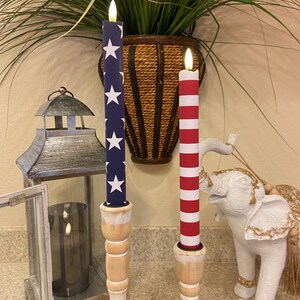 May include: Two decorative candles with a patriotic theme. One candle is navy blue with white stars, the other is red and white striped. Both are mounted on wooden candle holders. A metal lantern and a white elephant statue are also visible.