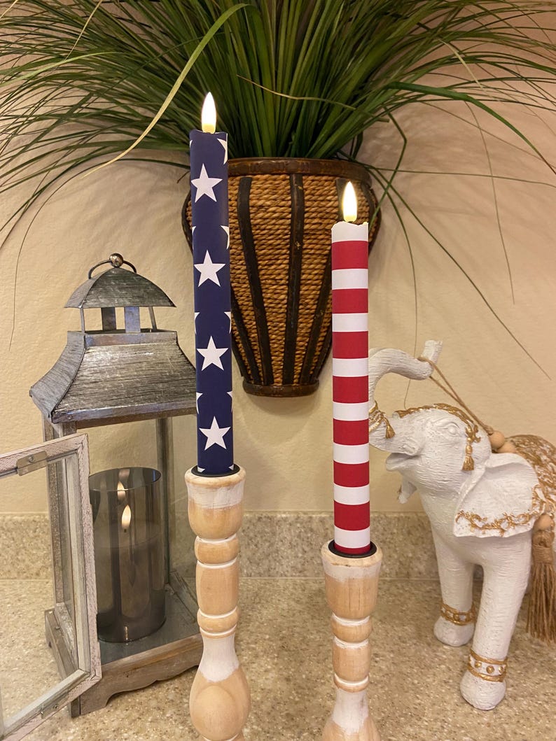 May include: Two decorative candles with a patriotic theme. One candle is navy blue with white stars, the other is red and white striped. Both are mounted on wooden candle holders. A metal lantern and a white elephant statue are also visible.