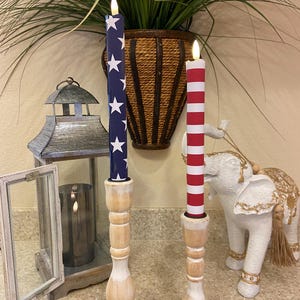 May include: Two decorative candles with a patriotic theme. One candle is navy blue with white stars, the other is red and white striped. Both candles are in wooden candle holders. A decorative lantern and elephant statue are in the background.