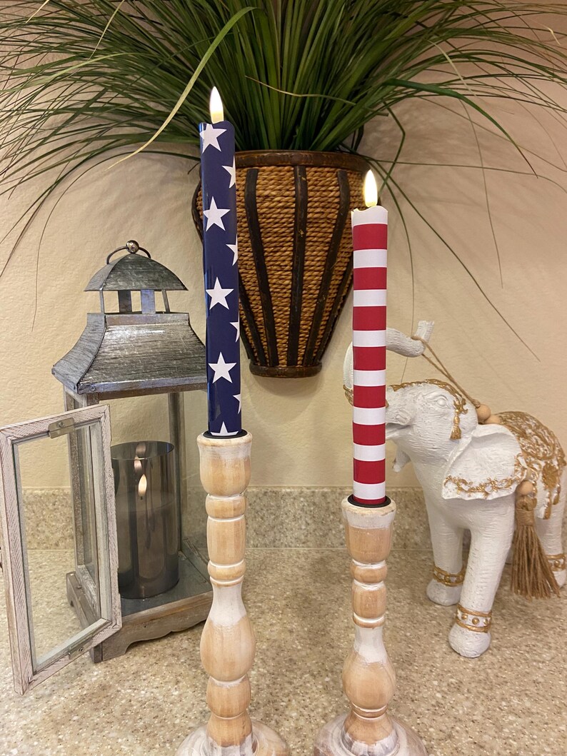 May include: Two decorative candles with a patriotic theme. One candle is navy blue with white stars, the other is red and white striped. Both candles are in wooden candle holders. A decorative lantern and elephant statue are in the background.