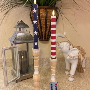 May include: Two decorative candles with patriotic designs. One candle has a blue background with white stars, the other red and white stripes. They are on wooden candle holders, with a remote control.