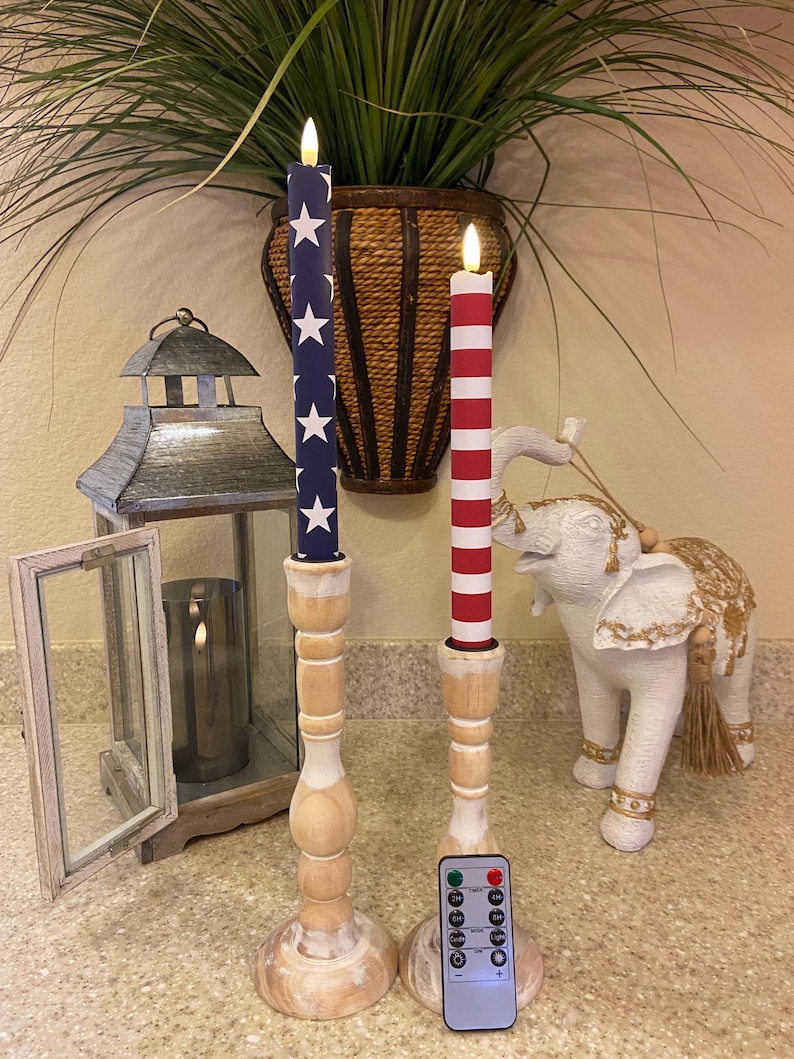 May include: Two decorative candles with patriotic designs. One candle has a blue background with white stars, the other red and white stripes. They are on wooden candle holders, with a remote control.