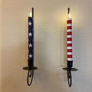 May include: Two wall-mounted candle holders, each with a battery-powered candle. One candle is navy blue with white stars, the other is red and white striped. Both have a flickering flame and are in black metal holders.