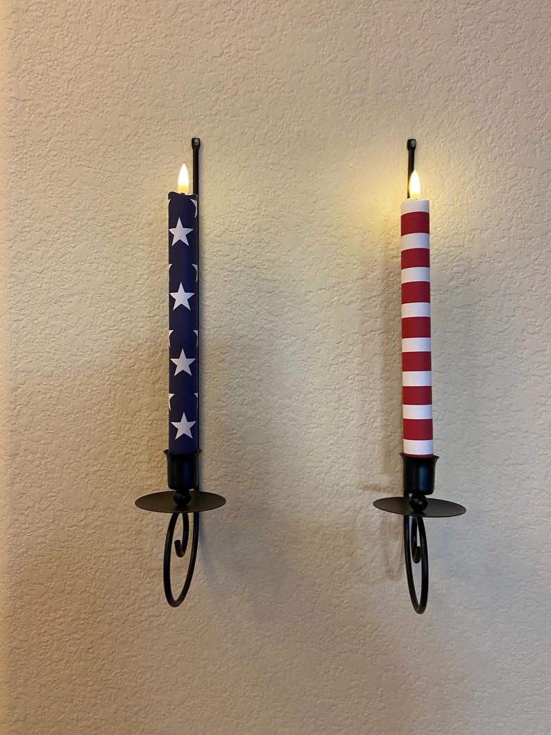 May include: Two wall-mounted candle holders, each with a battery-powered candle. One candle is navy blue with white stars, the other is red and white striped. Both have a flickering flame and are in black metal holders.