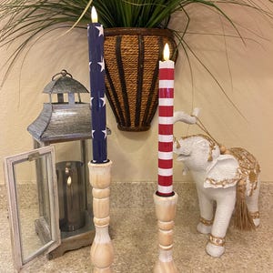 May include: Two decorative candles with wooden bases. One candle is navy blue with white stars, the other is red and white striped. The candles are in front of a plant and a decorative elephant statue.