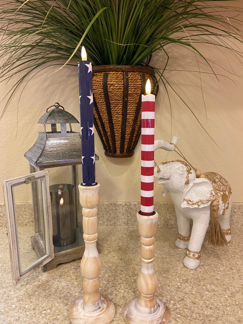 May include: Two decorative candles with wooden bases. One candle is navy blue with white stars, the other is red and white striped. The candles are in front of a plant and a decorative elephant statue.