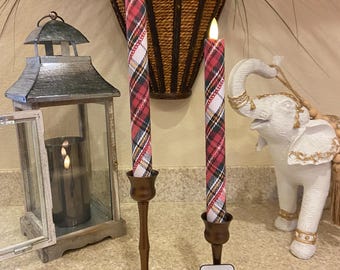 May include: Two battery-operated, plaid-patterned candles in bronze-colored candle holders. The candles have a red, white, and black plaid design. A remote control sits in front of the candles. A decorative lantern and elephant statue are in the background.