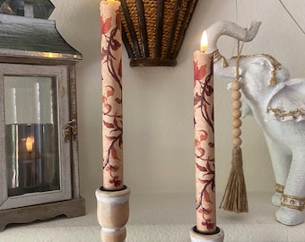 May include: Two tall, slender taper candles featuring a light beige background with a repeating pattern of dark red and brown autumn leaves and vines. Each candle is topped with a small, glowing LED flame and sits in a light-coloured wooden holder.