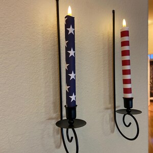 May include: Two wall-mounted candle holders with decorative candles. One candle is blue with white stars, the other is red and white striped. The holders are black metal with a curved design.