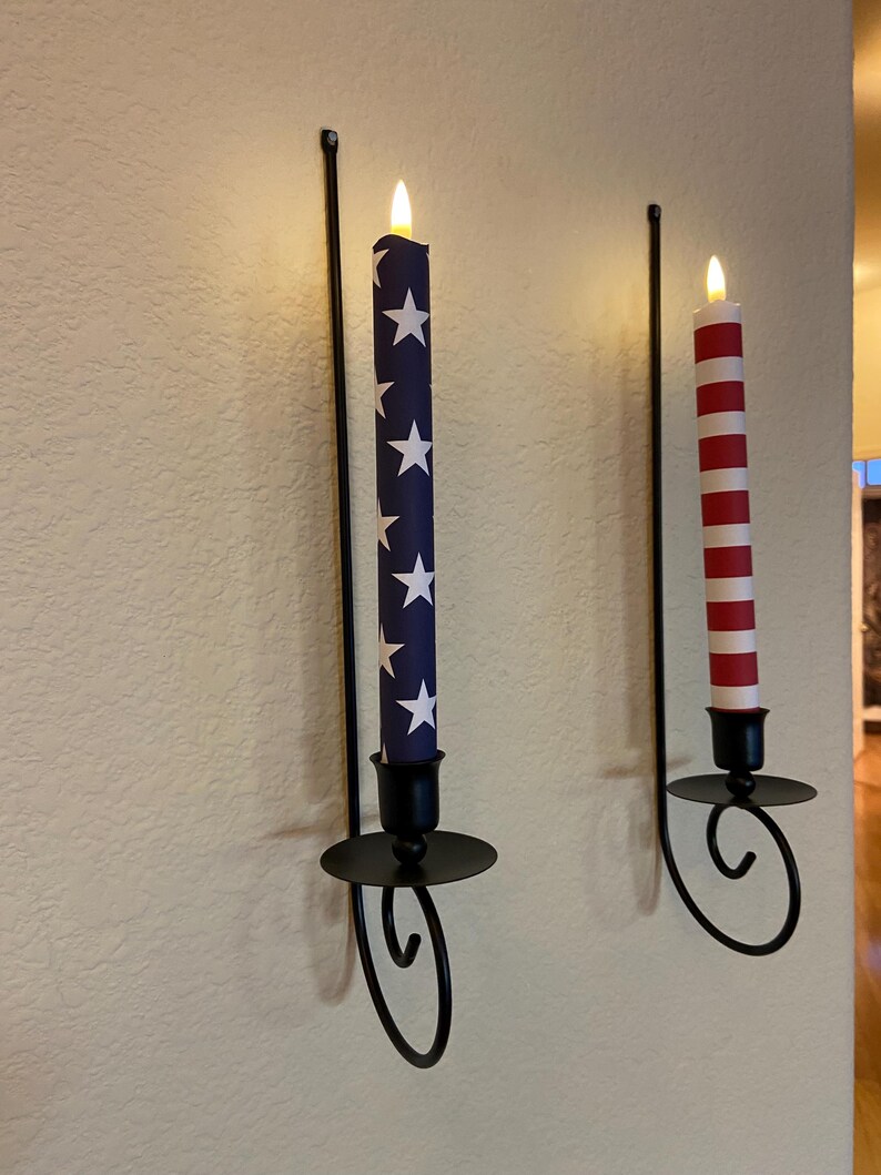 May include: Two wall-mounted candle holders with decorative candles. One candle is blue with white stars, the other is red and white striped. The holders are black metal with a curved design.