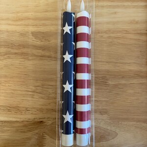 May include: Two LED candles in a clear plastic package. One candle is navy blue with white stars, the other has red and white stripes. The candles are designed to resemble the American flag. Each candle is approximately 20 cm tall.