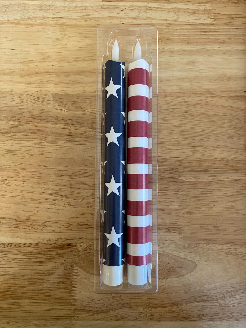 May include: Two LED candles in a clear plastic package. One candle is navy blue with white stars, the other has red and white stripes. The candles are designed to resemble the American flag. Each candle is approximately 20 cm tall.