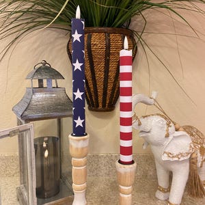 May include: Two decorative candles with a patriotic theme. One candle is navy blue with white stars, and the other has red and white stripes. Both are on wooden candle holders. A decorative lantern and elephant statue are also in the image.