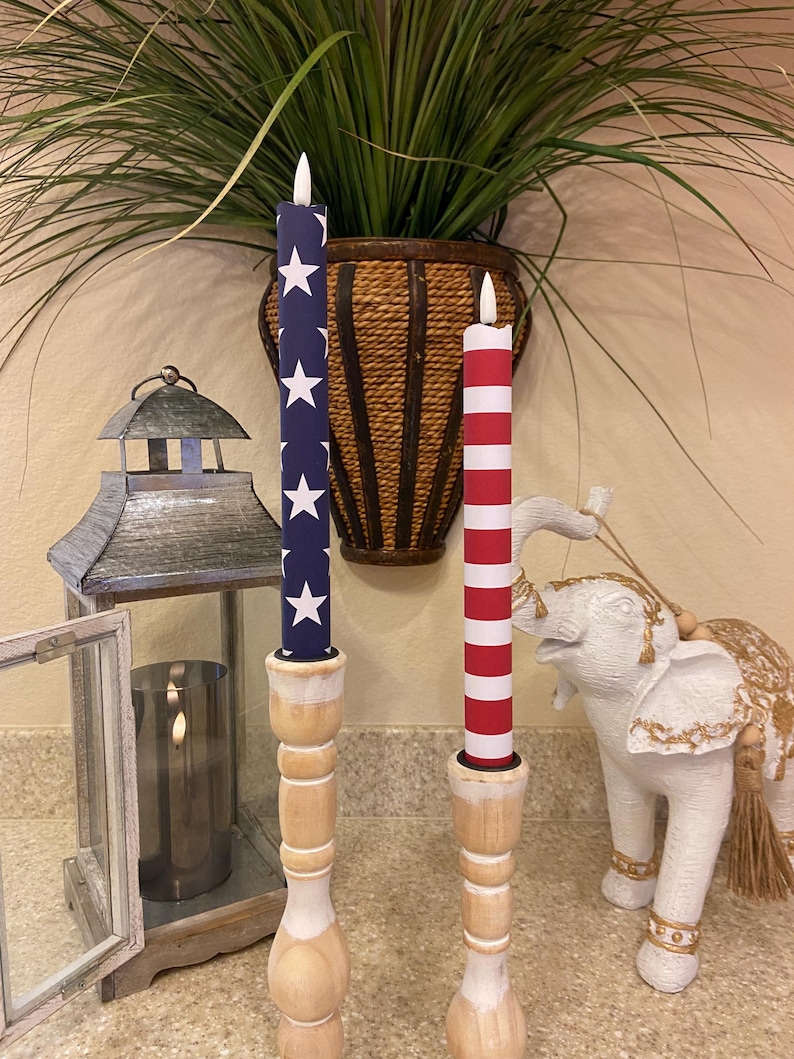 May include: Two decorative candles with a patriotic theme. One candle is navy blue with white stars, and the other has red and white stripes. Both are on wooden candle holders. A decorative lantern and elephant statue are also in the image.