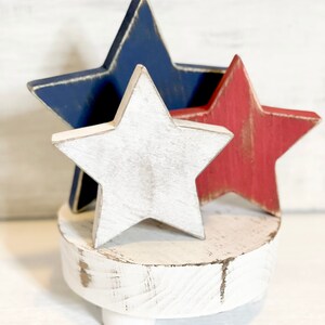 Puede incluir: Un pedestal de madera blanco con tres patas, coronado por tres estrellas de madera. Las estrellas est&aacute;n pintadas de rojo, blanco y azul.