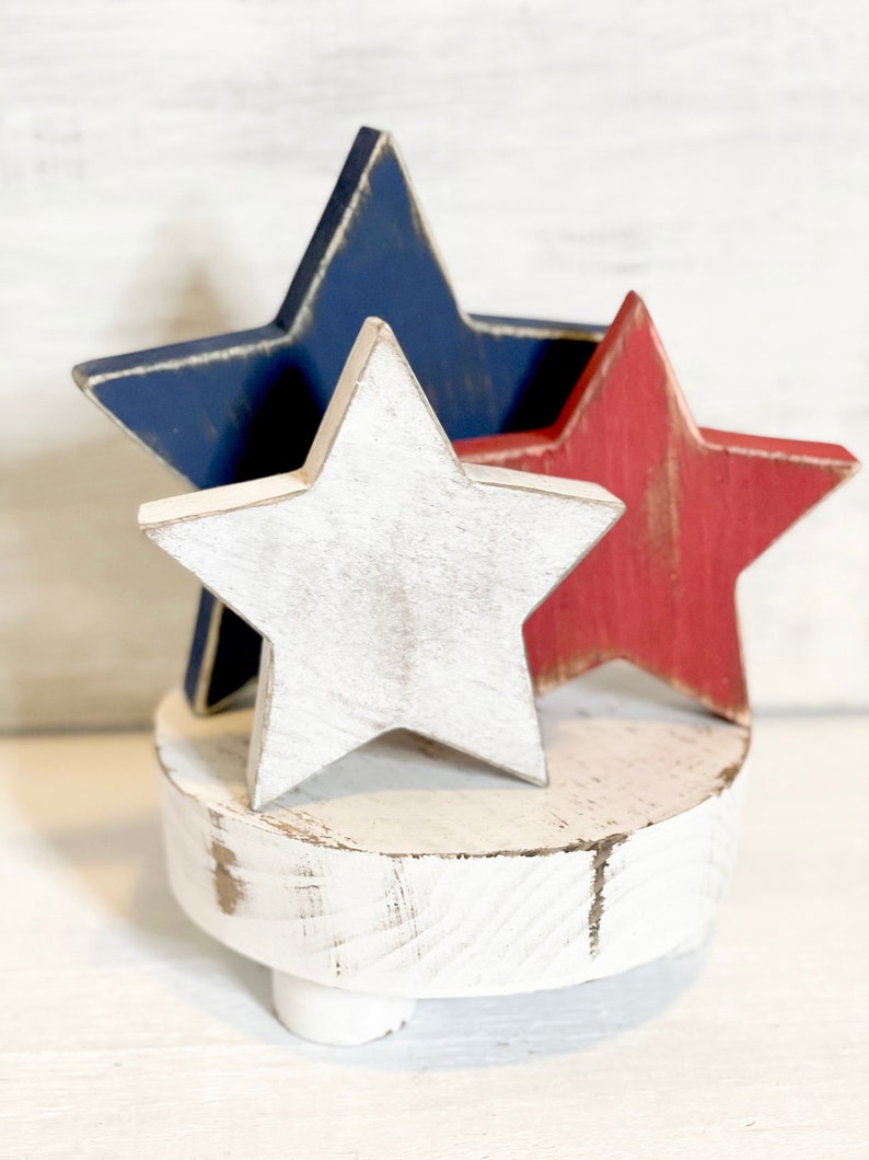 Puede incluir: Un pedestal de madera blanco con tres patas, coronado por tres estrellas de madera. Las estrellas est&aacute;n pintadas de rojo, blanco y azul.