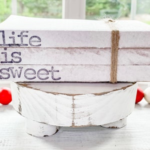 May include: White decorative books stacked on a white, distressed wooden pedestal. The books are tied with twine and feature the text "Life is Sweet" in black lettering. A string of red and white beads is in the foreground.