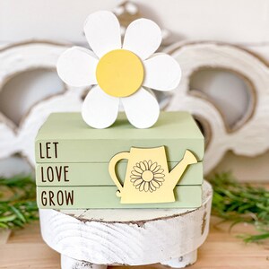 May include: A decorative wooden stack featuring the words "LET LOVE GROW" in brown lettering. A yellow watering can with a sunflower design is attached to the side. A large white and yellow daisy sits atop the stack.