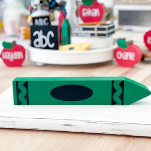 May include: A green wooden crayon with a black oval and decorative black accents. The crayon is on a white, distressed wooden surface. Other school-themed decorations, including red apple cutouts with names, are in the background.