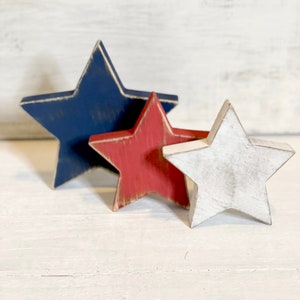 Puede incluir: Tres estrellas de madera en rojo, blanco y azul. Las estrellas est&aacute;n apiladas una encima de la otra, con la estrella azul en la parte inferior, la estrella roja en el medio y la estrella blanca en la parte superior.