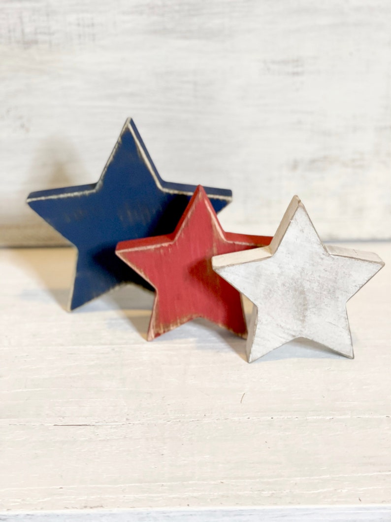 Puede incluir: Tres estrellas de madera en rojo, blanco y azul. Las estrellas est&aacute;n apiladas una encima de la otra, con la estrella azul en la parte inferior, la estrella roja en el medio y la estrella blanca en la parte superior.