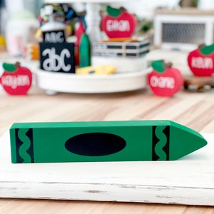 May include: A green wooden crayon decoration with a black oval and decorative black lines. The crayon is on a white wooden surface. Other school-themed decorations are in the background, including red apple cutouts with names.