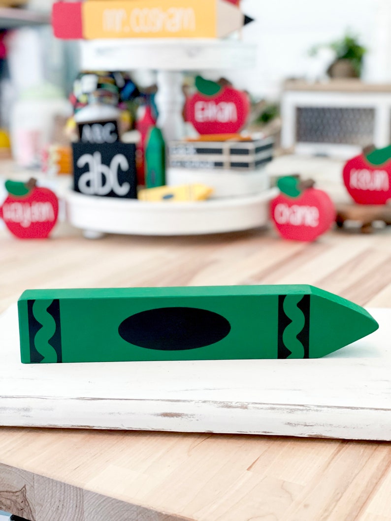 May include: A green wooden crayon decoration with a black oval and decorative black lines. The crayon is on a white wooden surface. Other school-themed decorations are in the background, including red apple cutouts with names.
