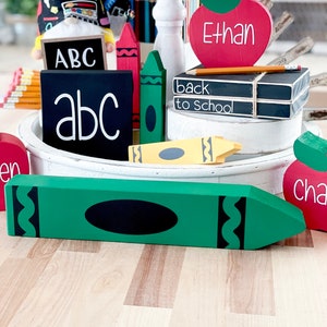May include: A collection of wooden school-themed decorations, including a large green crayon, red and green crayons, and apple-shaped signs with names. There are also blocks with the letters "ABC" and "back to school" written on them.