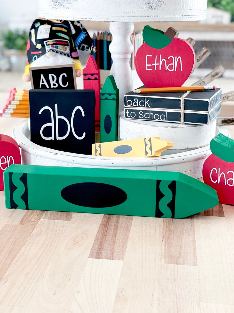 May include: A collection of wooden school-themed decorations, including a large green crayon, red and green crayons, and apple-shaped signs with names. There are also blocks with the letters "ABC" and "back to school" written on them.