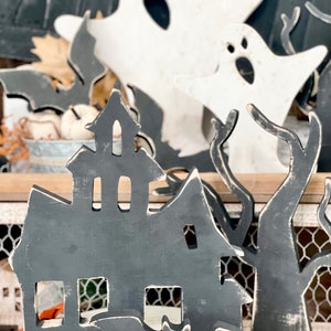 May include: Wooden Halloween decorations featuring a haunted house, bats, ghosts, and trees. The house and bats are painted dark gray with white accents, while the ghosts are white. The decorations are placed on a white surface with fall leaves.