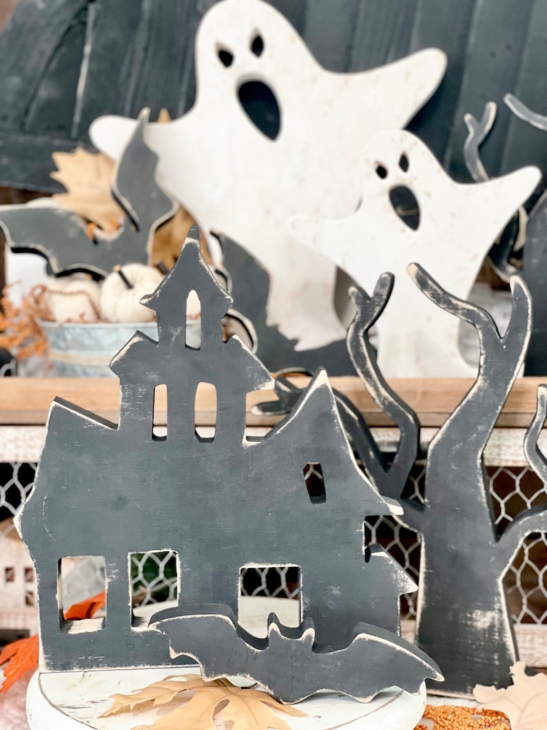 May include: Wooden Halloween decorations featuring a haunted house, bats, ghosts, and trees. The house and bats are painted dark gray with white accents, while the ghosts are white. The decorations are placed on a white surface with fall leaves.