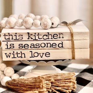 May include: A stack of three white books with the text "this kitchen is seasoned with love" printed on them. The books are tied together with twine and are sitting on a white pedestal with a beaded garland draped around the base.