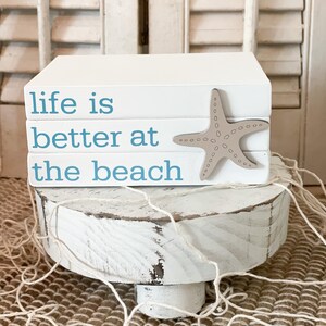 May include: White wooden block sign with the text "life is better at the beach" in blue lettering. A tan starfish is placed on top of the sign. The sign is sitting on a white wooden pedestal.