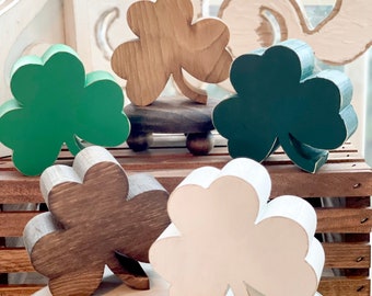 May include: Five wooden shamrock shapes in various colors, including green, brown, and white. The shamrocks are arranged on a white surface.