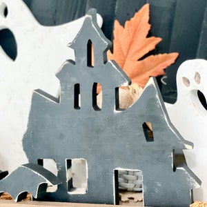May include: A gray wooden haunted house decoration with a distressed finish, featuring cut-out windows and a bat silhouette. The Halloween-themed decor includes a white ghost and an orange leaf.