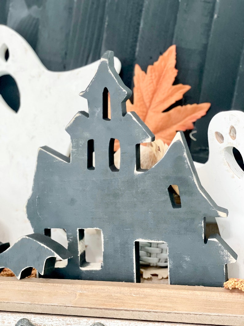 May include: A gray wooden haunted house decoration with a distressed finish, featuring cut-out windows and a bat silhouette. The Halloween-themed decor includes a white ghost and an orange leaf.