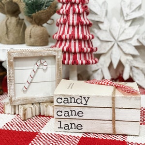 May include: A stack of three white books with the words "candy cane lane" printed on them. The books are tied together with twine and a red and white checkered ribbon. The books are sitting on a red and white checkered tablecloth.