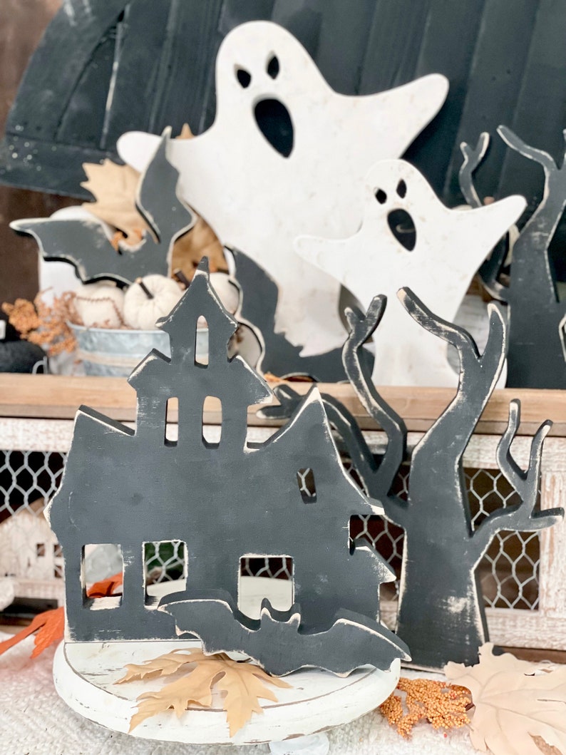 May include: Halloween-themed wooden decorations. The image features a black haunted house, bats, and trees, along with white ghost cutouts. The decorations are placed on a white surface with fall leaves, creating a spooky seasonal display.