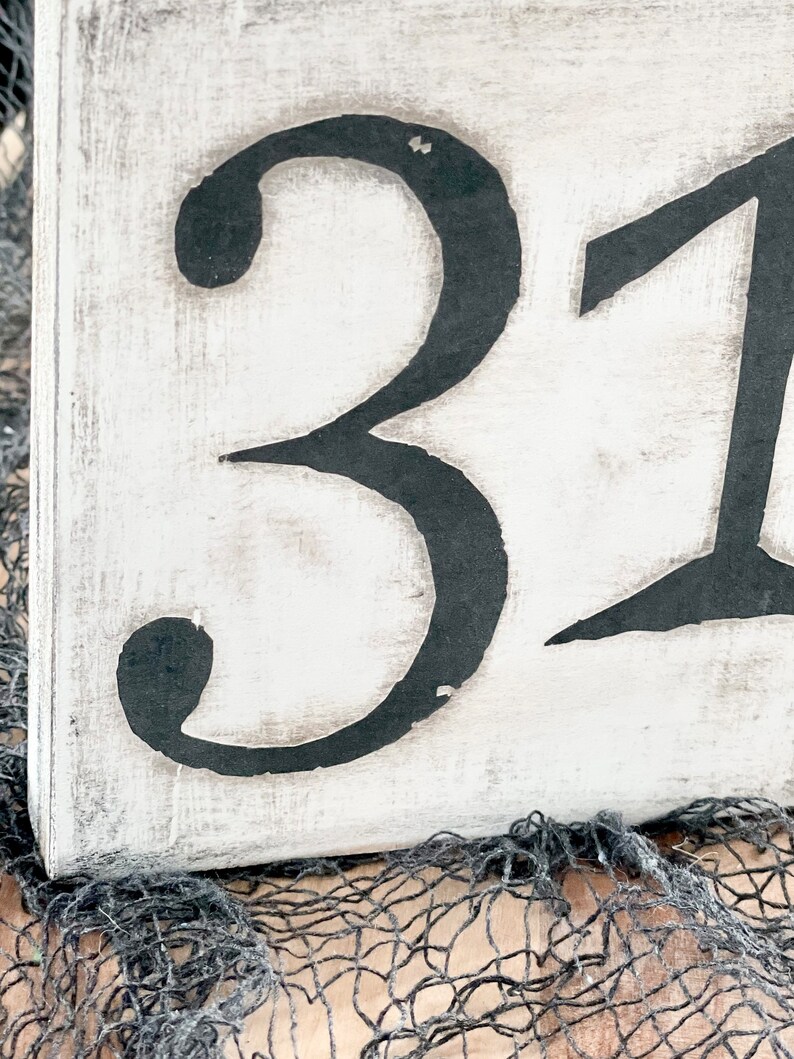 May include: A white wooden sign with black painted numbers, "31", on a distressed background. The sign is sitting on a black and brown mesh surface.