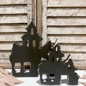May include: Two dark gray wooden haunted house decorations with white trim. The houses have cut-out windows and doors. The background features a white shuttered window and fall leaves.