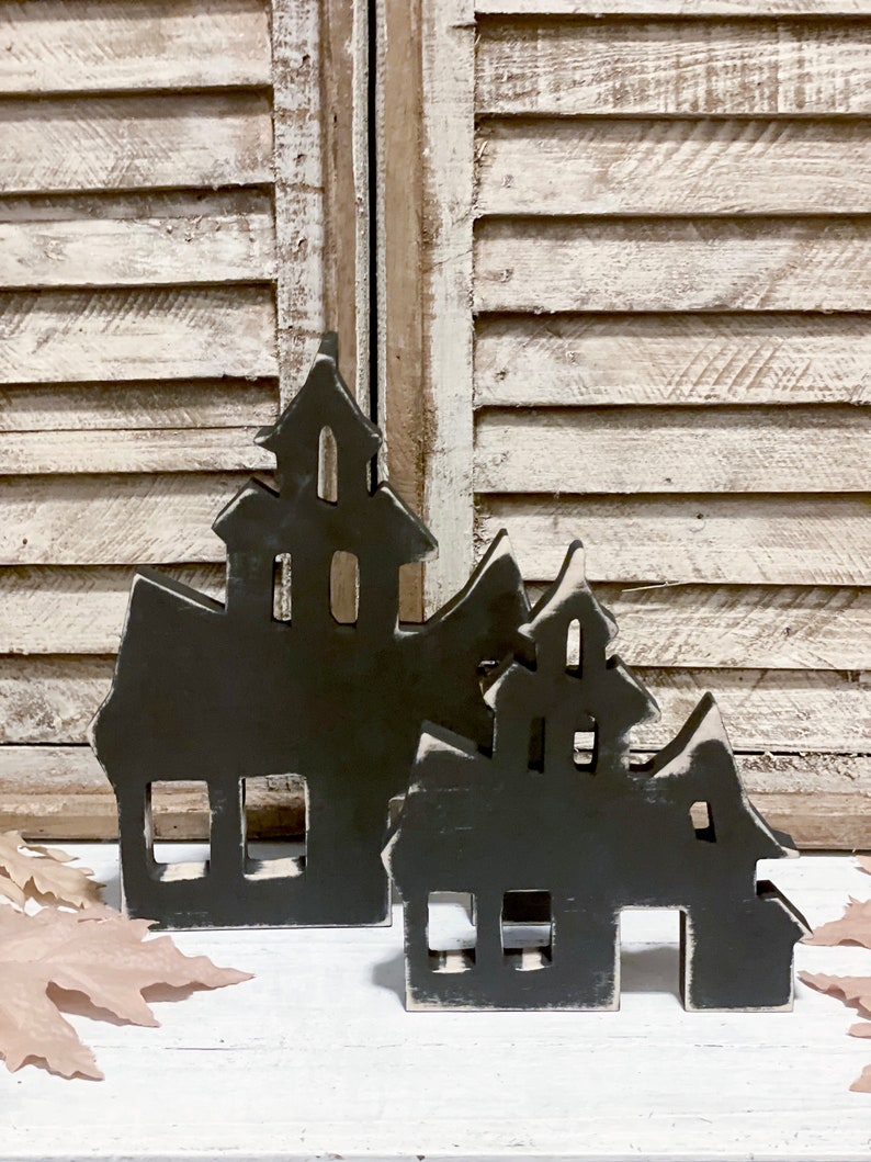 May include: Two dark gray wooden haunted house decorations with white trim. The houses have cut-out windows and doors. The background features a white shuttered window and fall leaves.