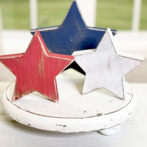 Puede incluir: Tres estrellas de madera en colores rojo, blanco y azul sobre un pedestal redondo blanco. Las estrellas est&aacute;n desgastadas y tienen un aspecto r&uacute;stico.