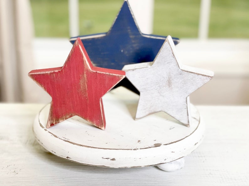 Puede incluir: Tres estrellas de madera en colores rojo, blanco y azul sobre un pedestal redondo blanco. Las estrellas est&aacute;n desgastadas y tienen un aspecto r&uacute;stico.