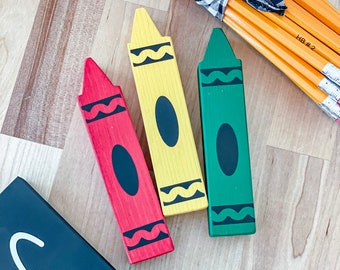 May include: Three wooden crayons, one red, one yellow, and one green, with black wavy lines and a black circle on each. The crayons are on a wooden surface with a bundle of yellow pencils tied with a black and white checkered ribbon.