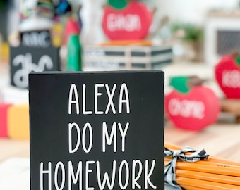 May include: Black chalkboard sign with white text that reads "Alexa do my homework".